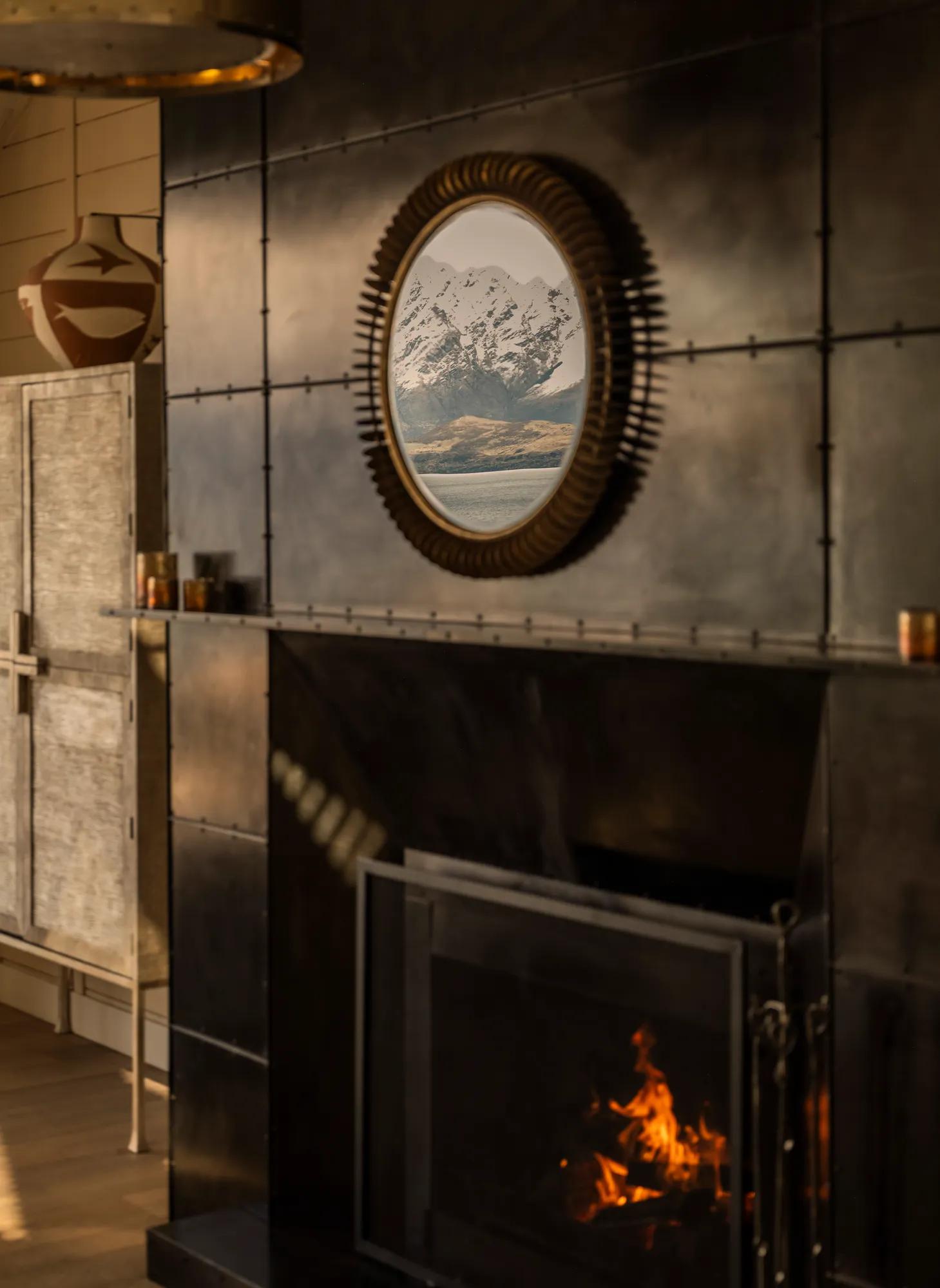 Steel surround open fire in the living room of a luxury villa, in the mirror above the fire are reflections of a lake and snow-covered mountains. 