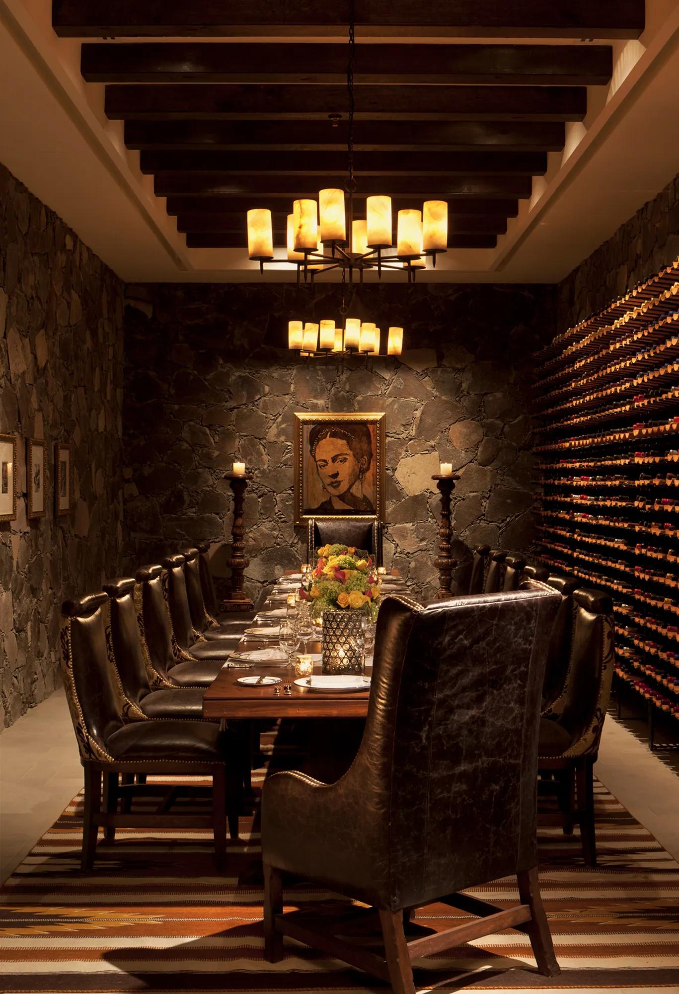 Long wooden table set for 14 guests inside a wine cellar, surrounded by wine shelves and soft lighting, with a framed artwork on the wall.