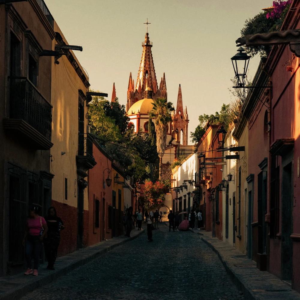Cobblestone street bathed in warm sunset light, lined with colorful red, orange, and yellow houses, with the Parroquia de San Miguel Arcángel visible in the distance.