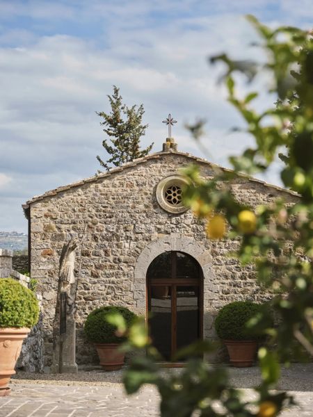 Una storica cappella in pietra con portale ad arco e croce, circondata da piante in fiore.