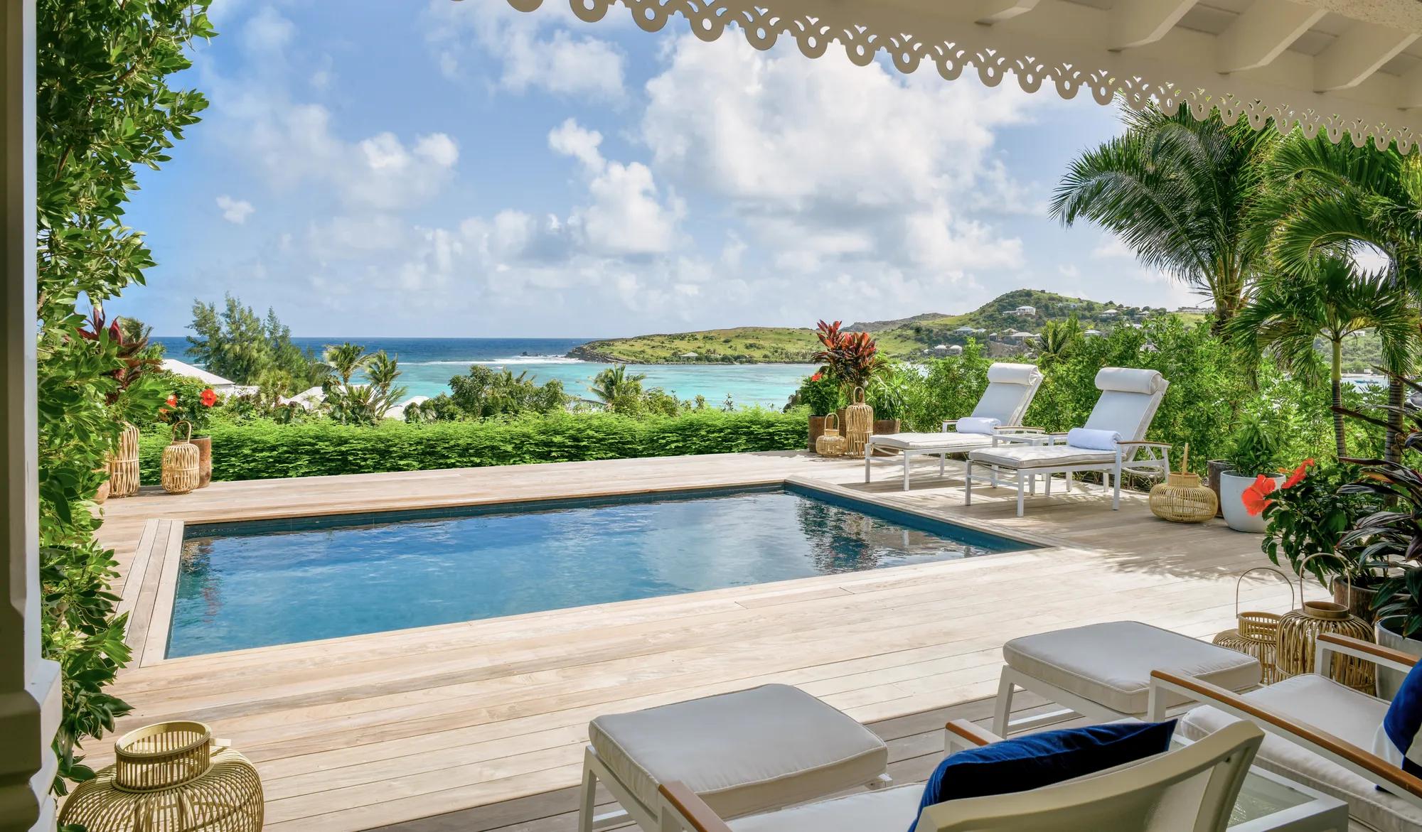 A pool in a St. Barth resort, surrounded by greenery and lounge chairs, overlooking the ocean during the day.