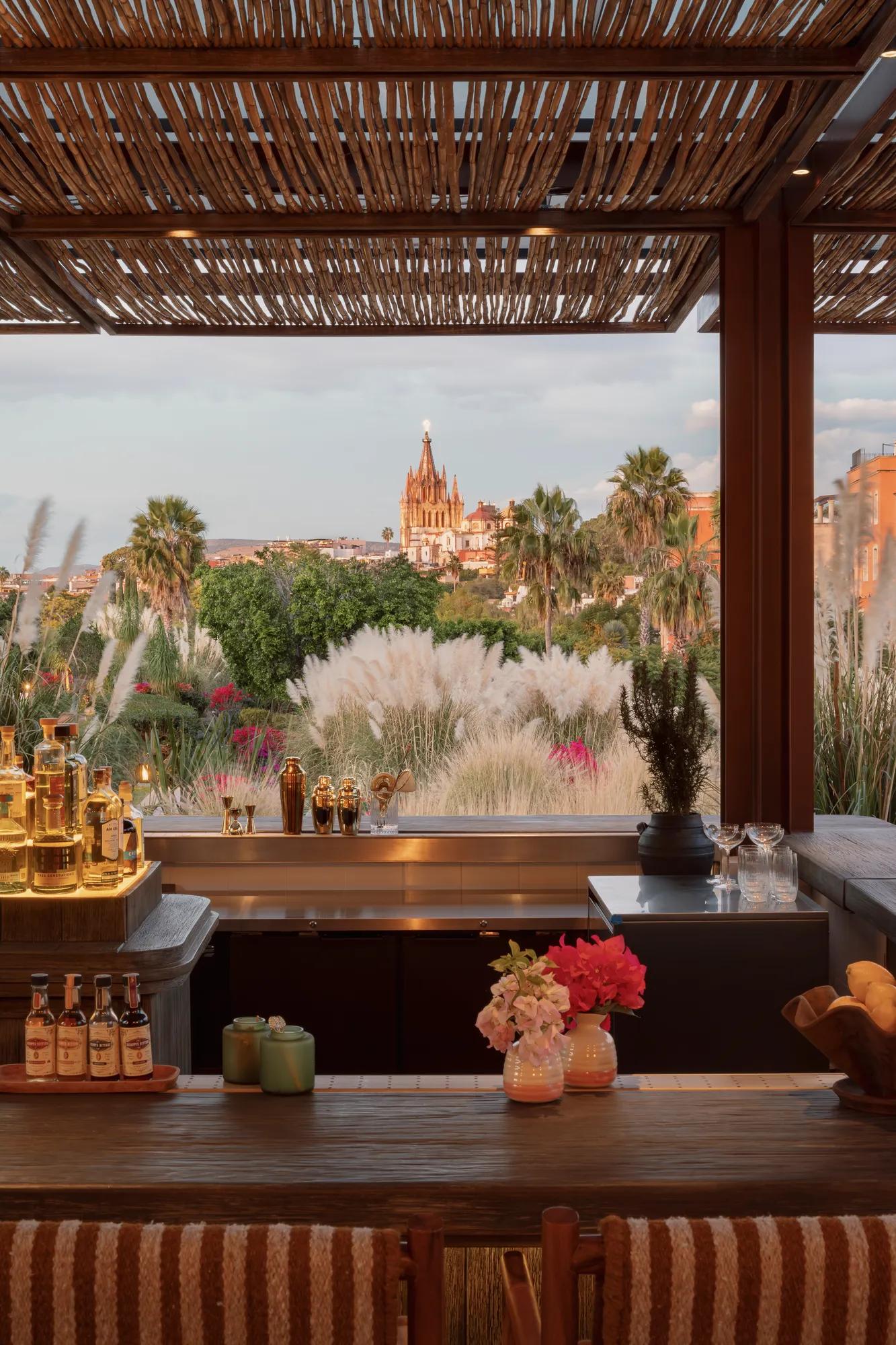 An open-air bar with bottles displayed on a backlit shelf, overlooking the hotel gardens and the Parroquia in the distance. 