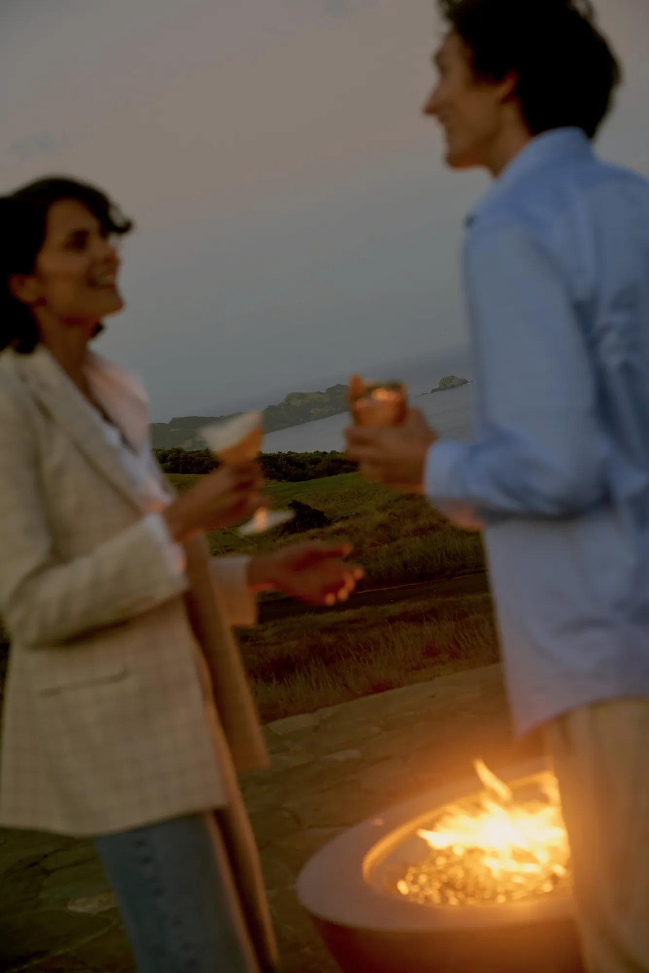Two people chat over cocktails next to the fire pit at Rosewood Kauri Cliffs, behind is the golf course, ocean and Cavalli Islands. 