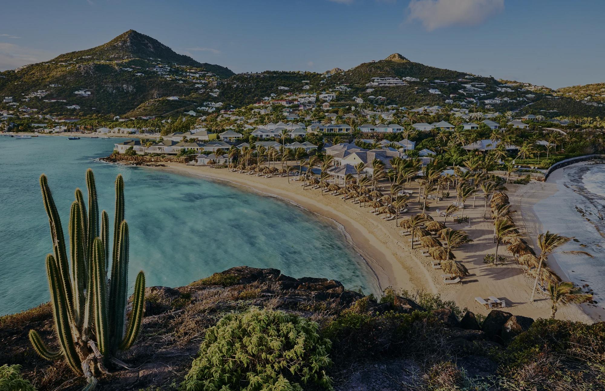 St. Barth Beach with clear water, resort   buildings, a mountain, and thatched   umbrellas along the beach. 
