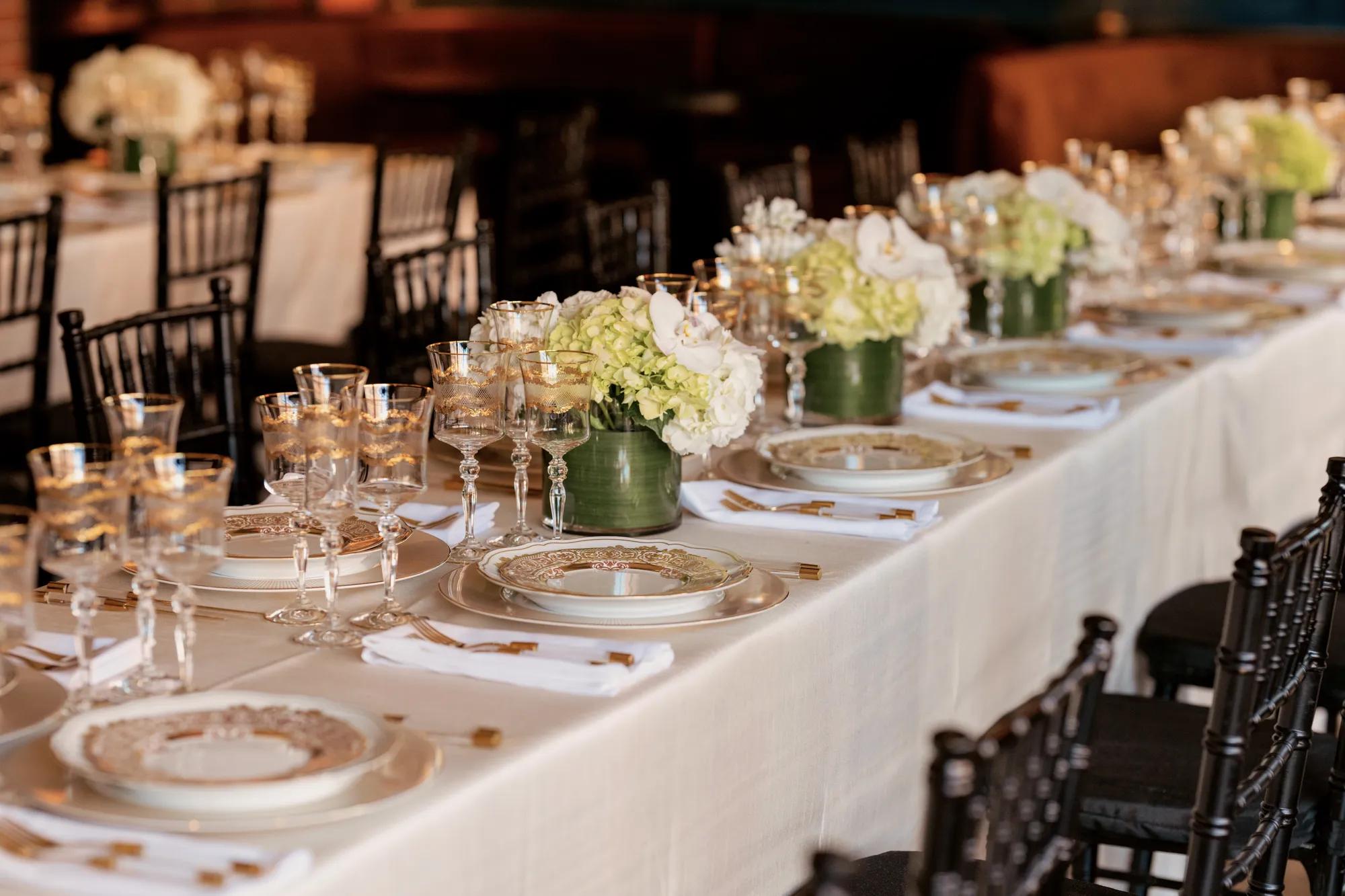 View of a long table setting in the CUT Main Dining Room featuring an ivory linen and gold trimmed flatware, plateware, and glassware with black chairs in view.