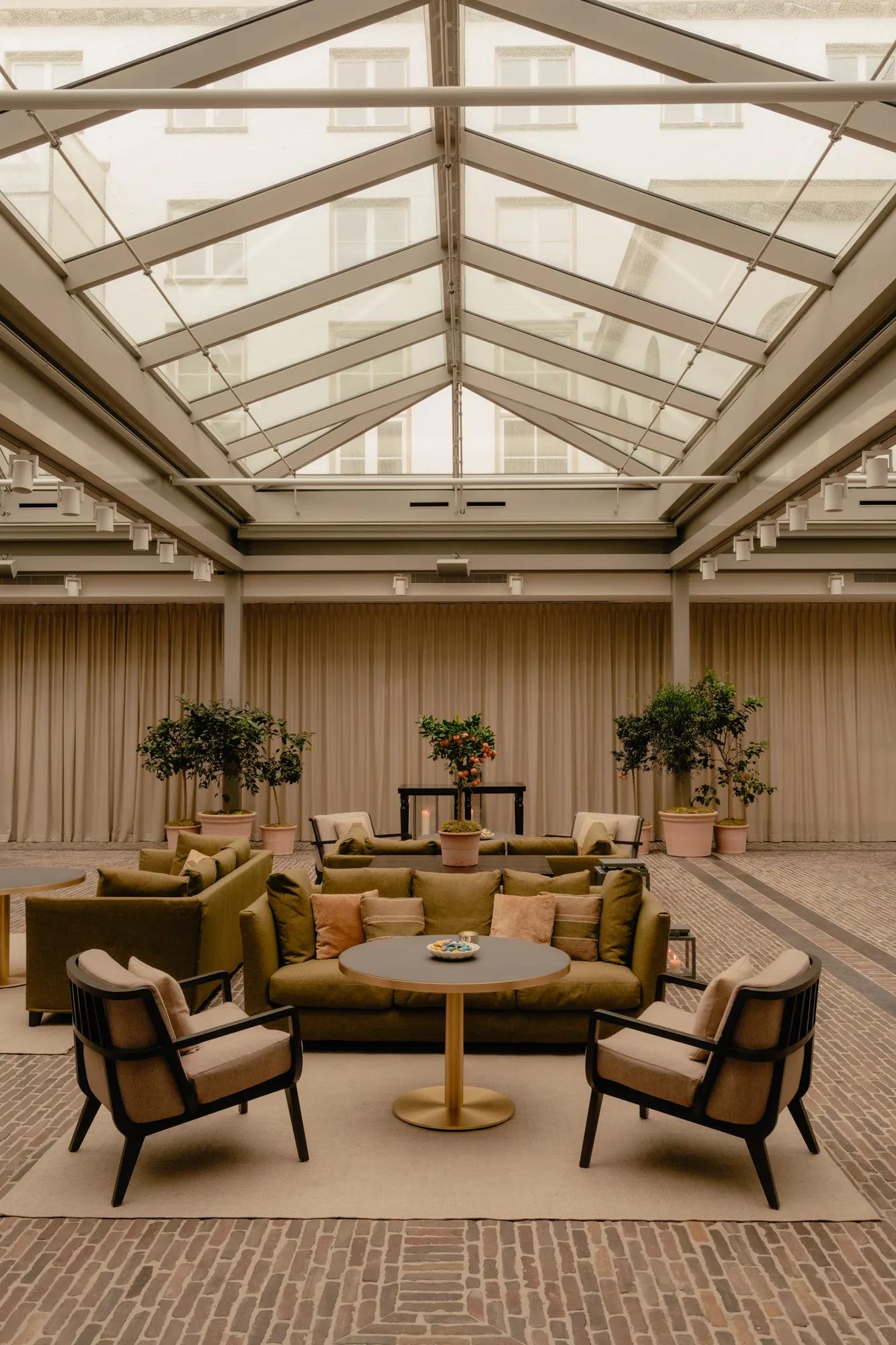 A glass covered courtyard with brick floors, sofas, chairs, tables and potted plants. 