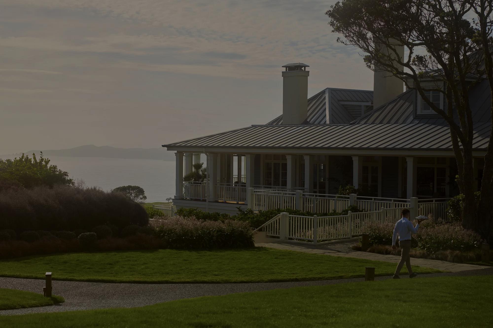 Waiter carrying a room service tray along a path back toward the main lodge at Rosewood Kauri Cliffs, in the distance you can see the ocean and the Bay of Islands.