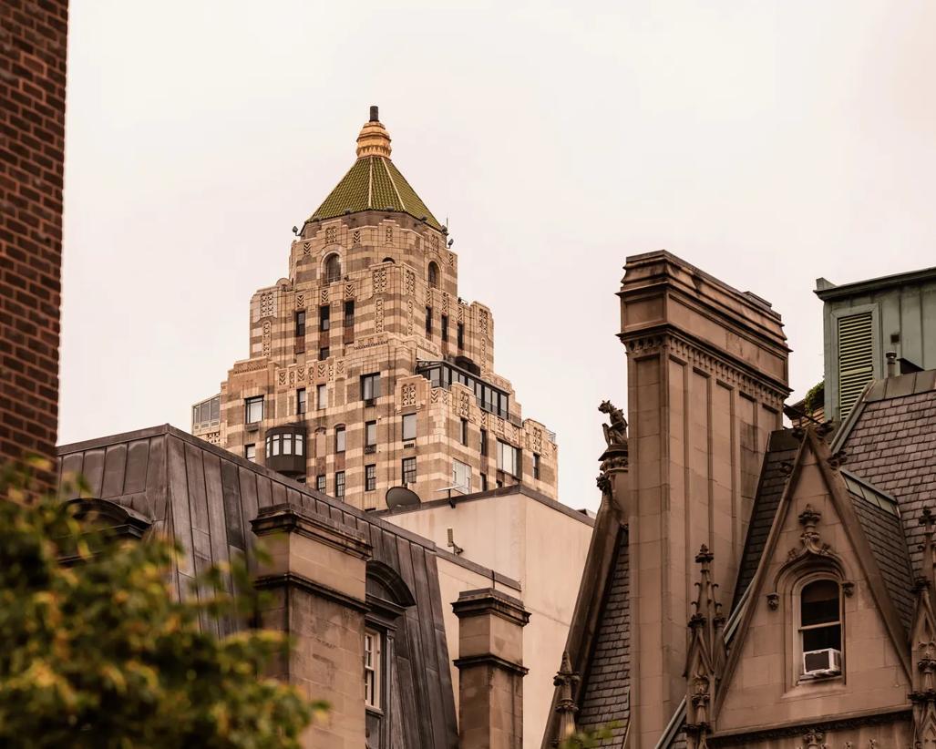 View of the iconic tower peekng through a layered cityscape of hostoric rooftops on the Upper East Side of Manhattan and the lush green trees of Central Park. 