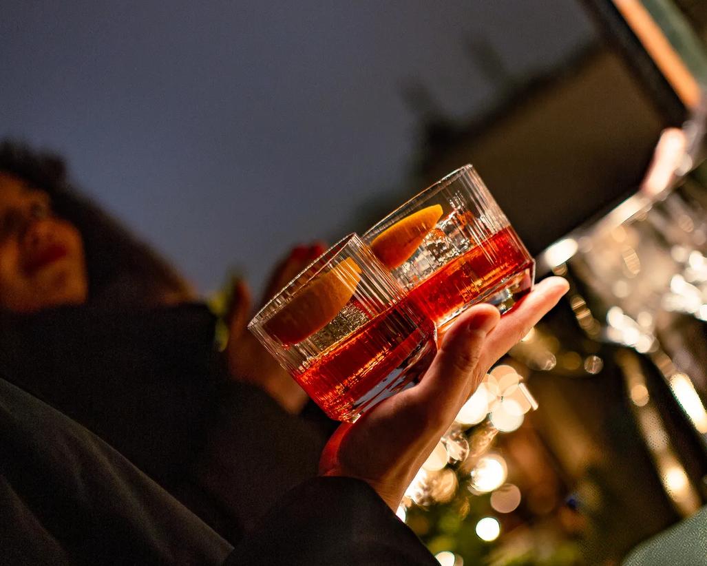 Hand holding two cocktails with orange garnishes at Würstl & Sprudl on the Rosewood Vienna rooftop.