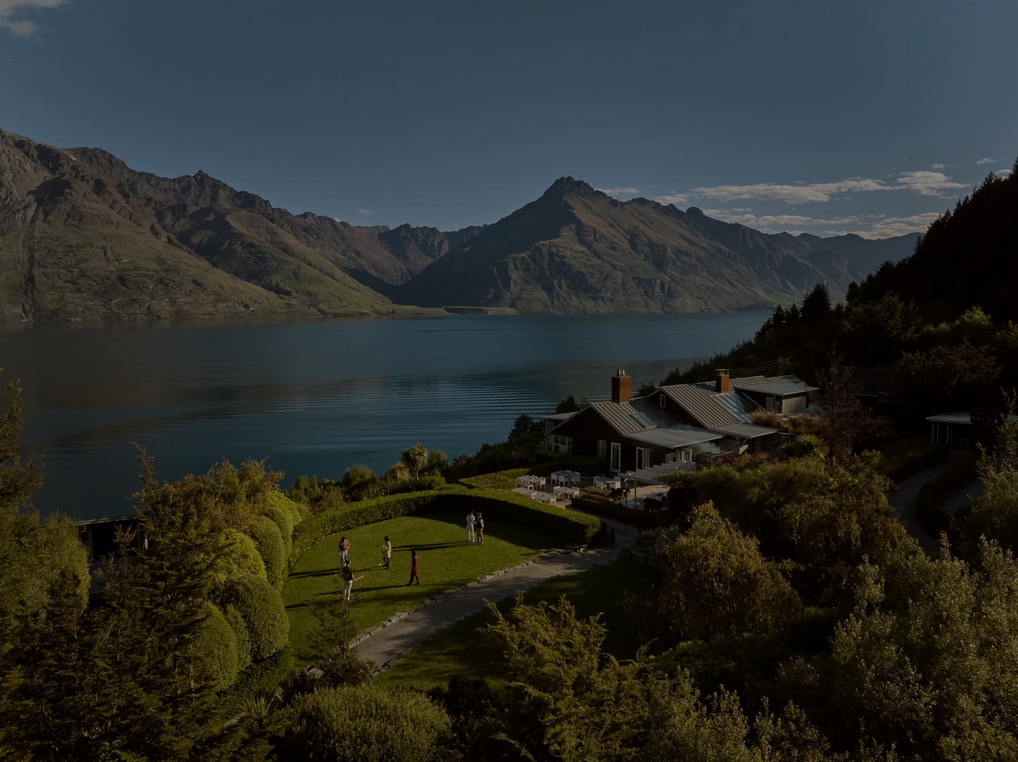 Aerial image of guests playing pétanque on the lush green lawn with the main lodge behind, in the distance is Lake Wakatipu and the surrounding mountain ranges.