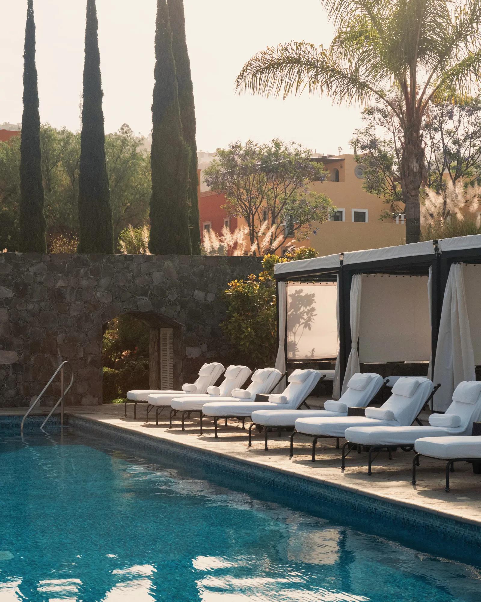 View of the pool area at Rosewood San Miguel de Allende with lounge chairs in the foreground, cabanas along the edge, pine trees and palm trees behind, and the exterior of residences further in the background.