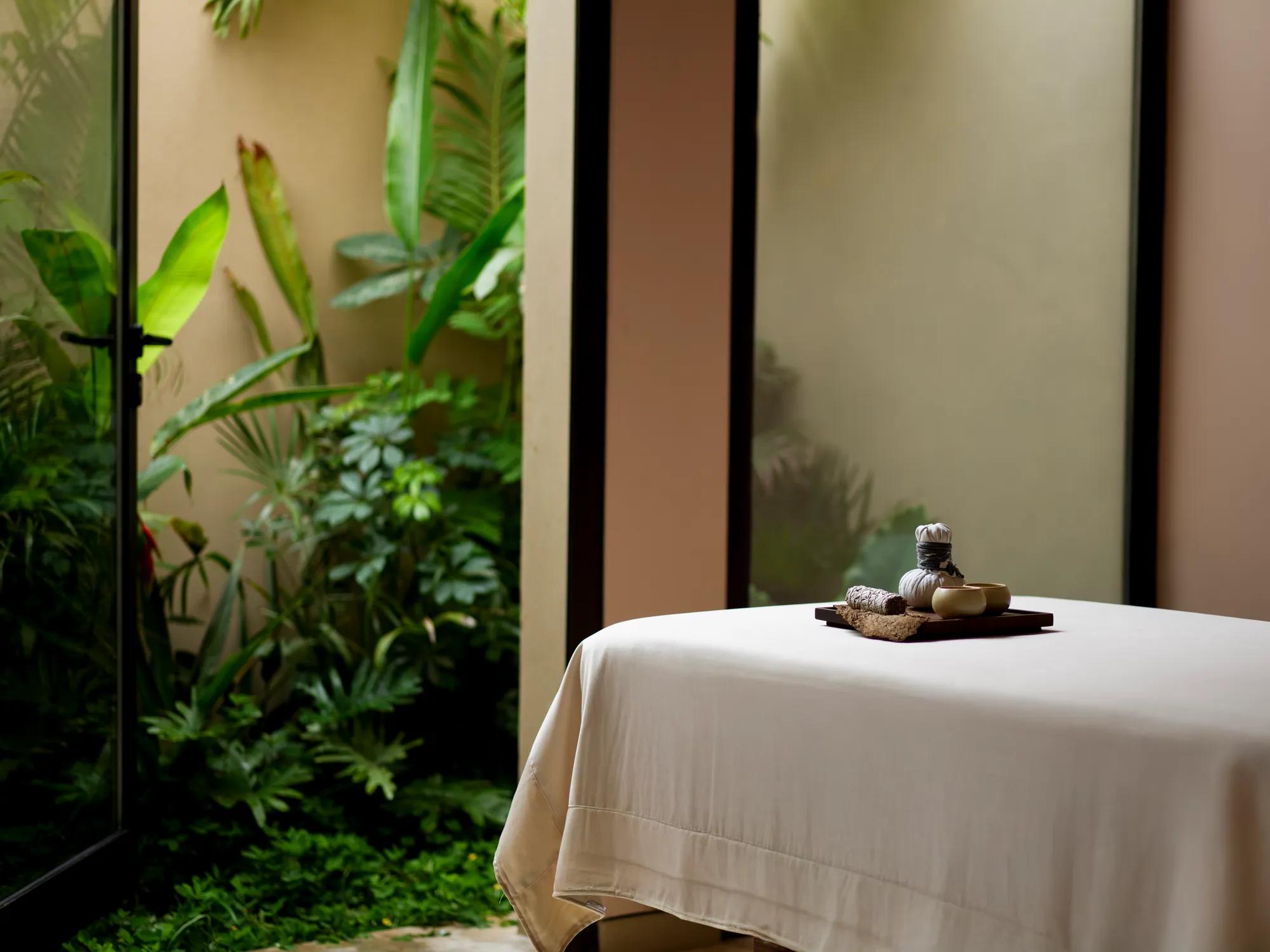 Spa massage table with white linens beside a window overlooking lush tropical greenery.