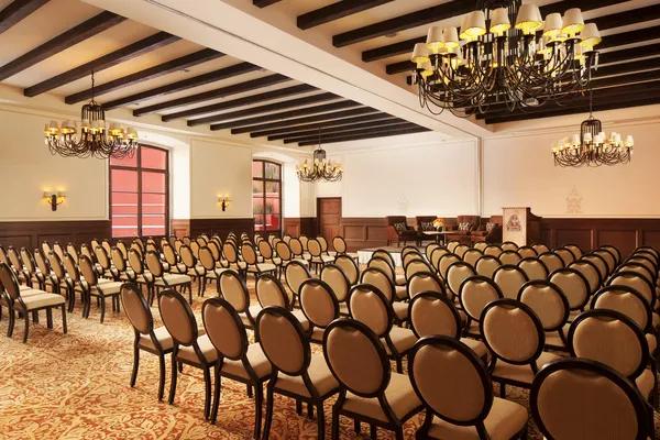 Ballroom set in auditorium style with multiple rows of chairs facing a presentation area, featuring carpeted floors, curtained windows, and warm lighting.