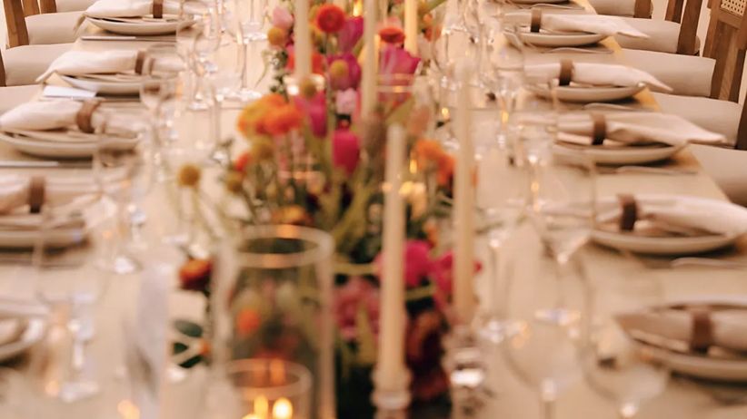 Linear view of a pink-toned wedding table setup with the ocean as a backdrop, soft hanging lights above, and palm trees framing the scene.