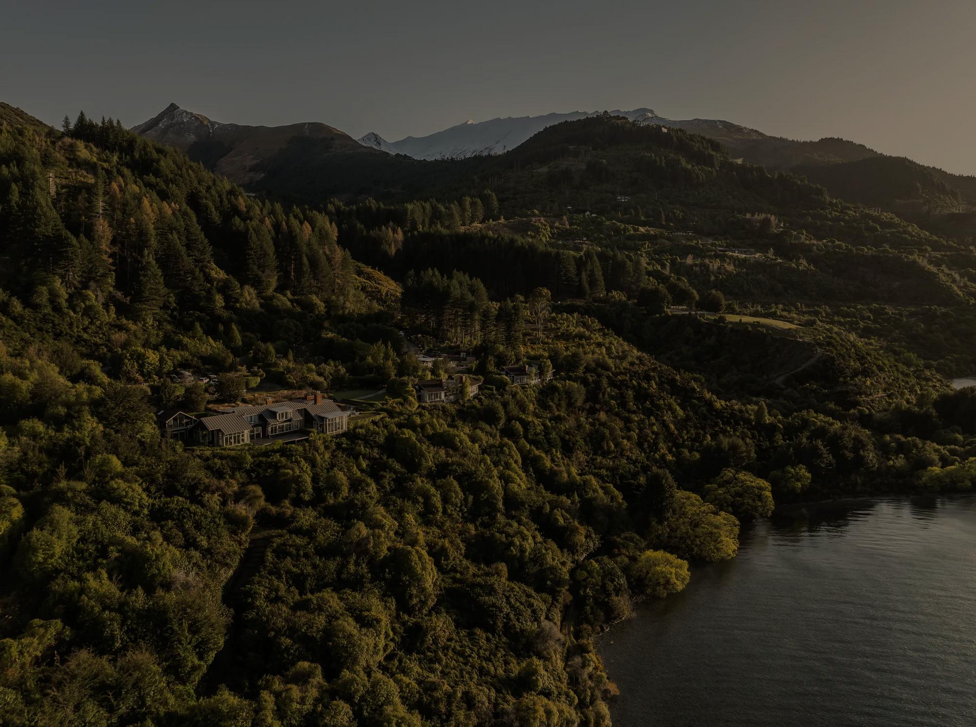 Aerial taken from the lake looking back at Rosewood Matakauri nestled into the surrounding native bush with snow-capped mountains behind. 