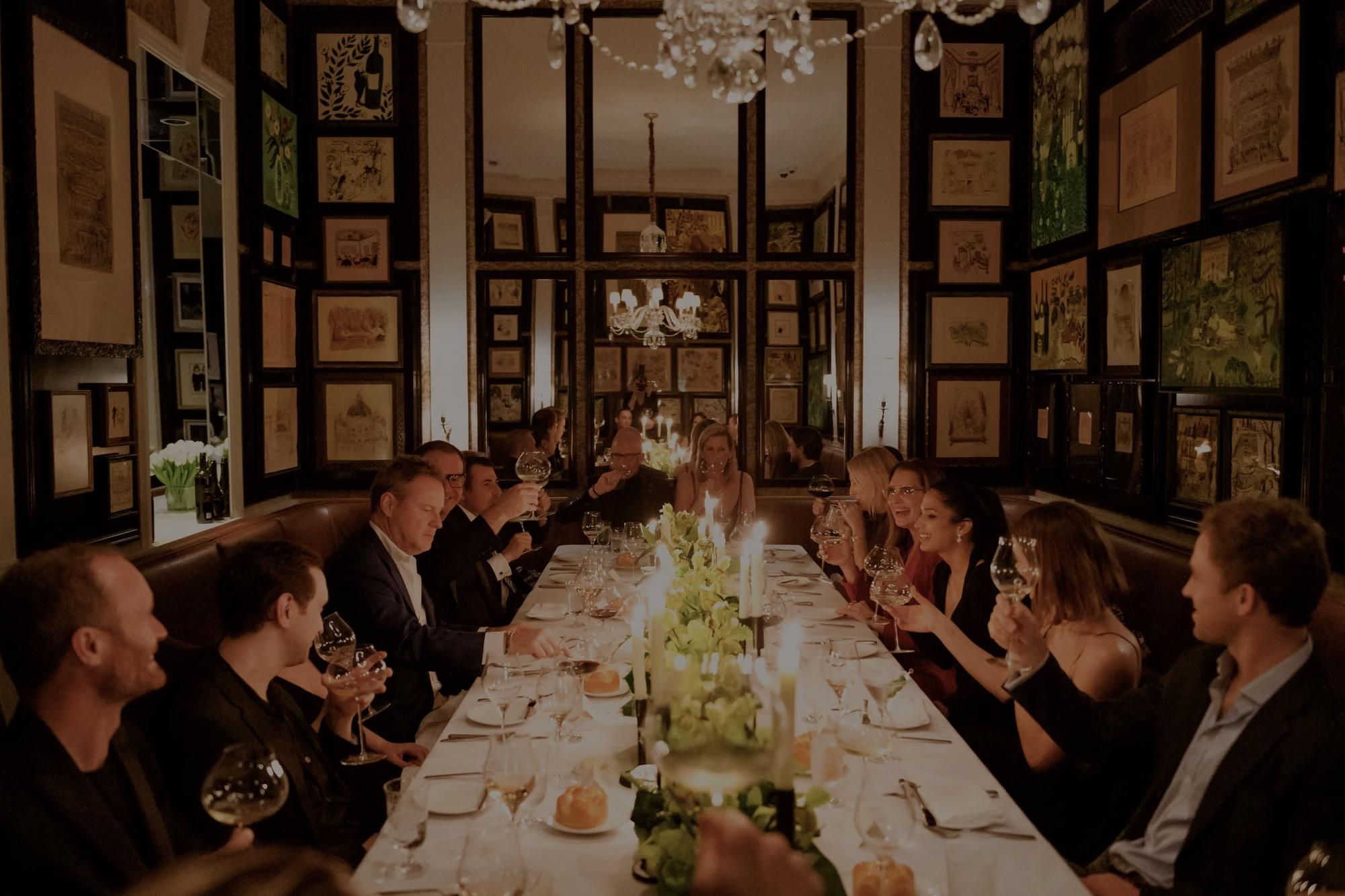 A lively dinner party is held in the sem-private room at Dowling's at The Carlyle, featuring a long, elegantly set table surrouded by well-dressed guest raising wine glasses in a toast. The wals are decorated with framed artwork, and a chandelier hangs abpve, casting a celebratory scene. 