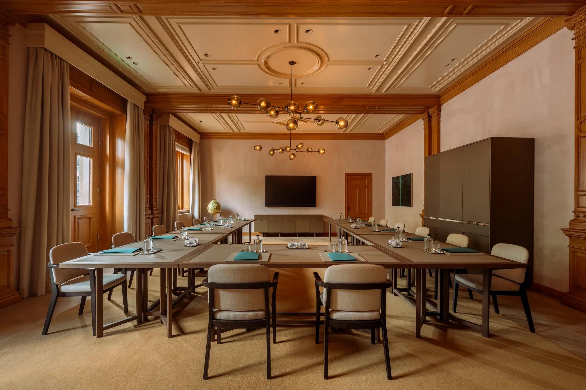 A meeting room with boardroom set up with traditional wooden paneling and floors, panelled ceilings and two chandeliers. 