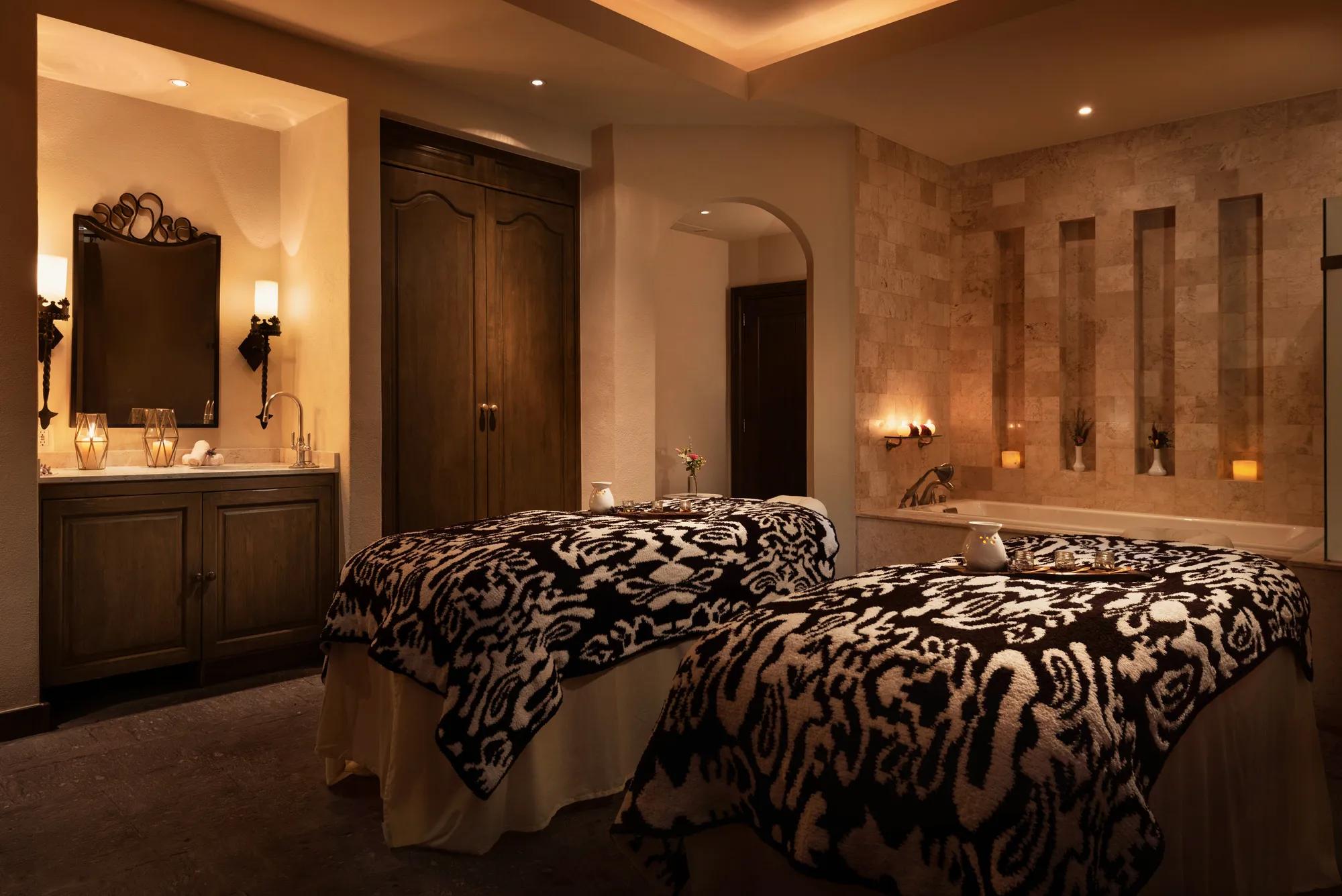 Two spa beds with embroidered blankets, a small table with a pitcher between them, and a bathtub in the background. On the left side, a sink with a mirror, two wall lamps, and candles create a warm, serene ambiance. A small closet stands beside the vanity.