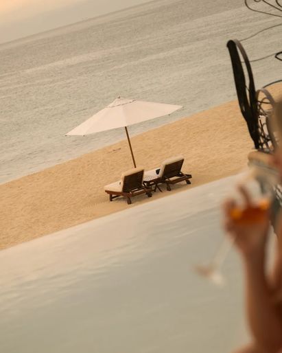 Blurred woman enjoying a drink beside an infinity pool overlooking a quiet beach with lounge chairs and an umbrella at sunset in Los Cabos.