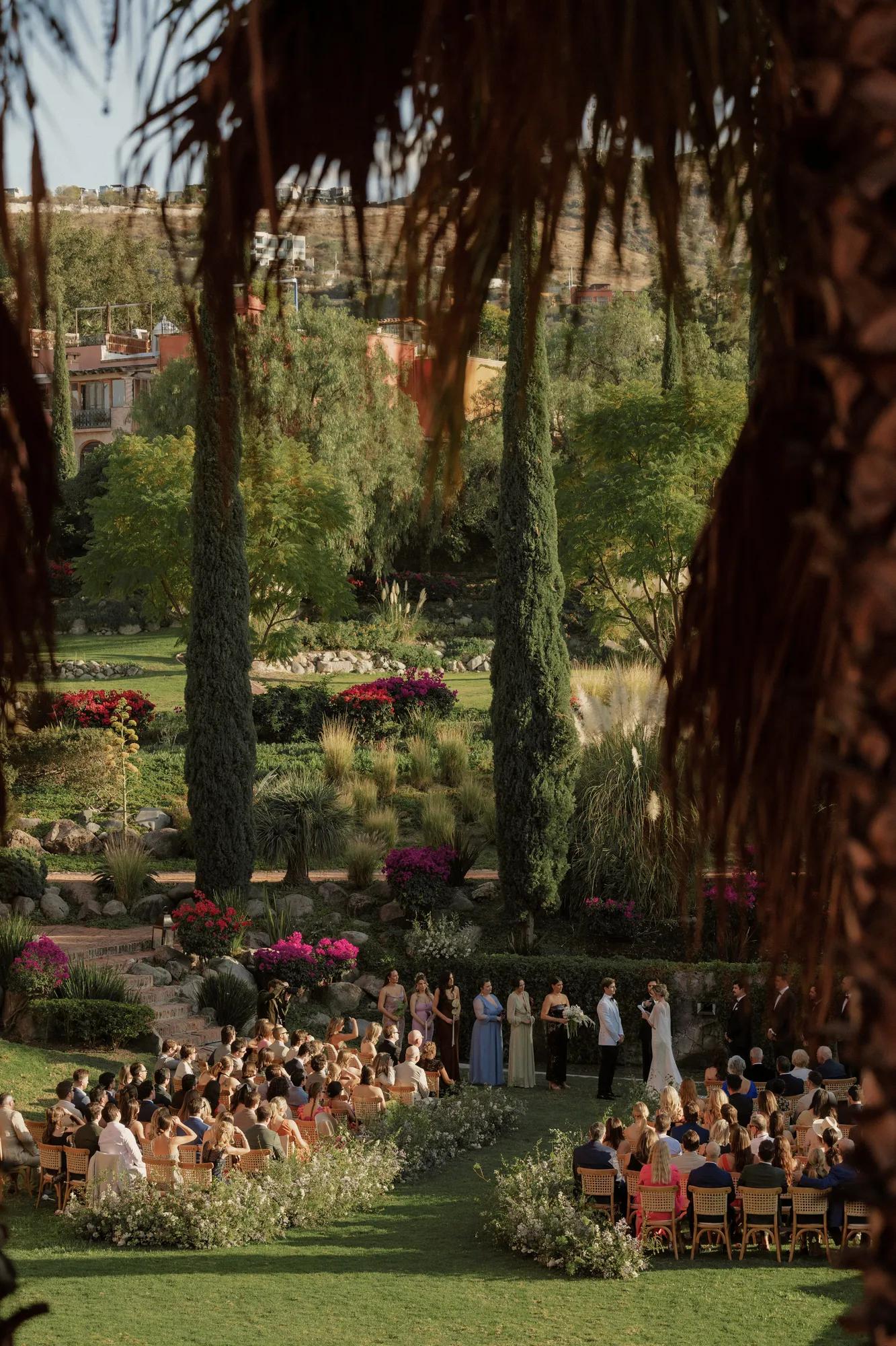 Bride and groom facing each other during a garden wedding ceremony, surrounded by bridesmaids, seated guests, and lush greenery.