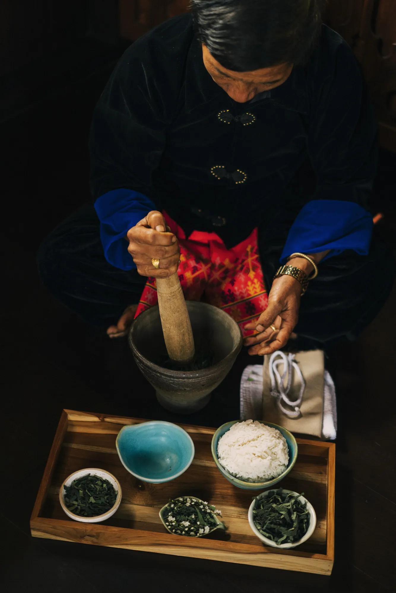 A woman wearing a traditional robe grinding herbs with a mortar and pestle.