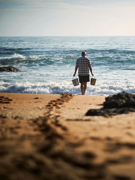 Male walking on the beach to the ocean with two metal buckets to collect water to make salt.