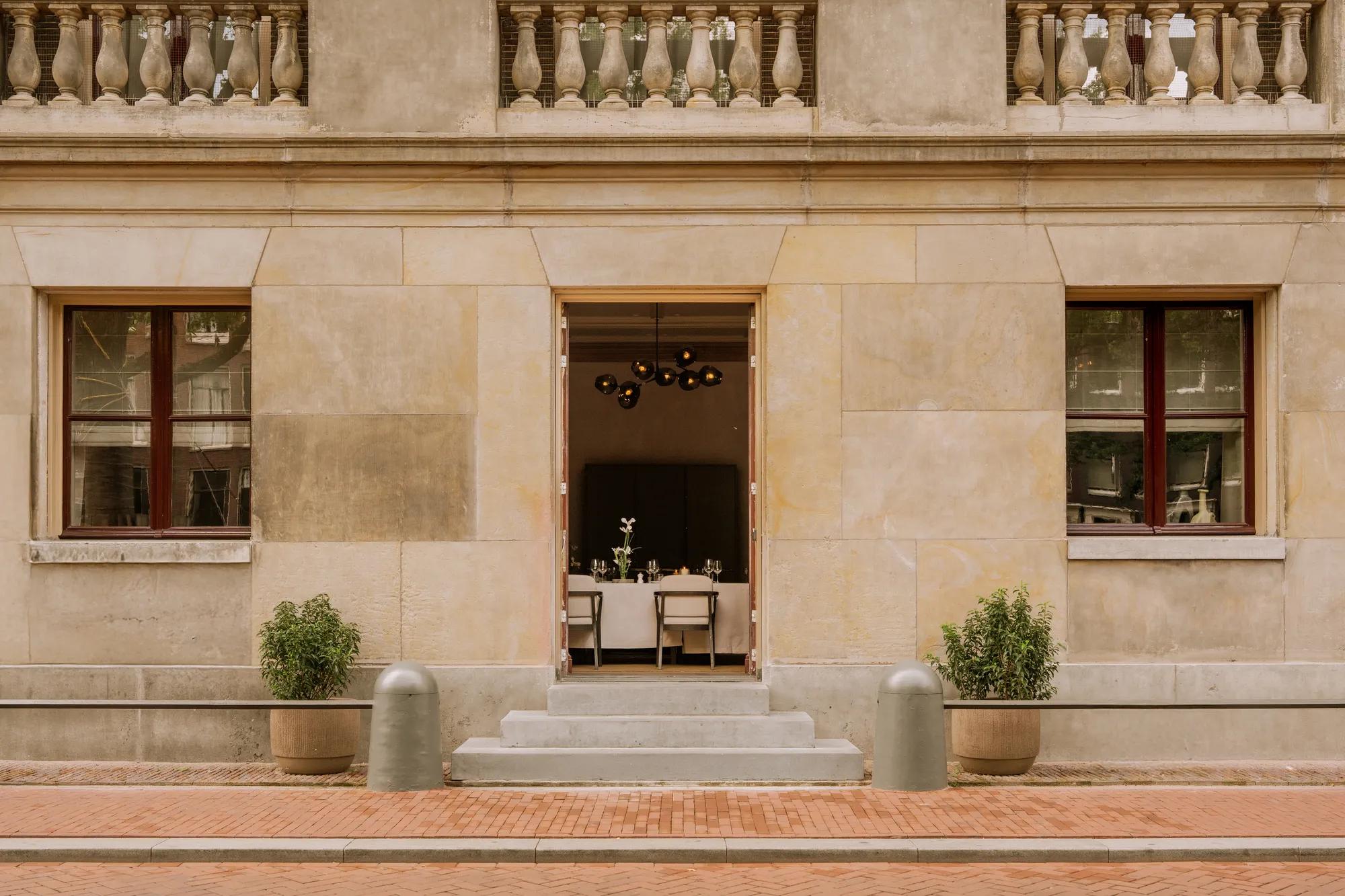 An immense stone facade with steps leading to an open door and two windows, showing a meeting room set up a tables in white table cloths. 