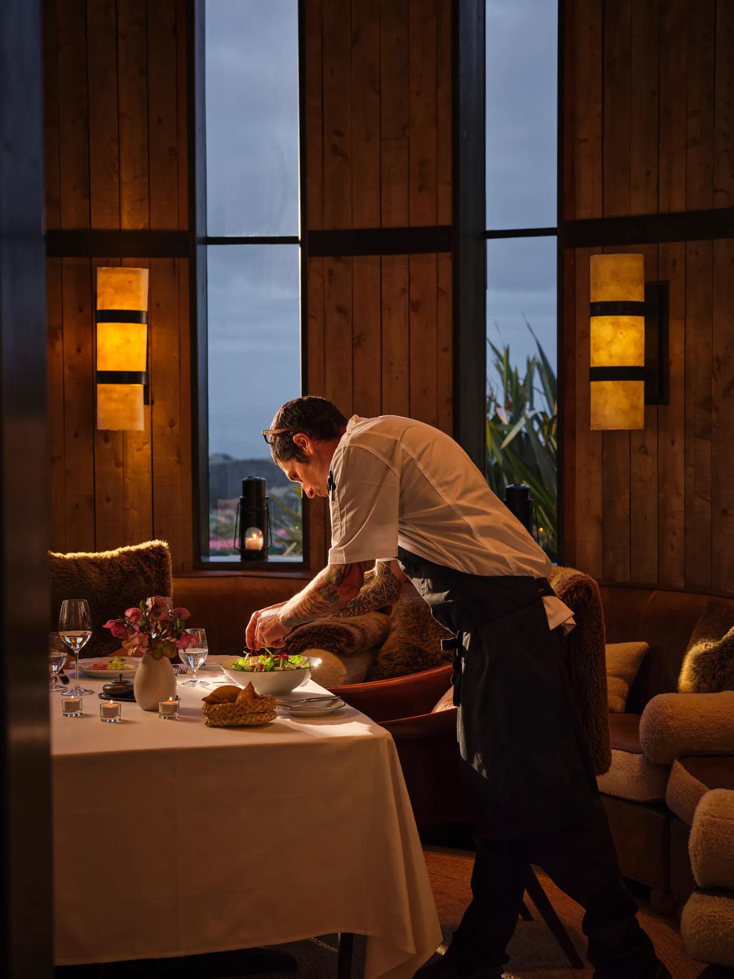 Chef preparing a dish at a dimly lit table with mountain views through floor-to-ceiling windows.