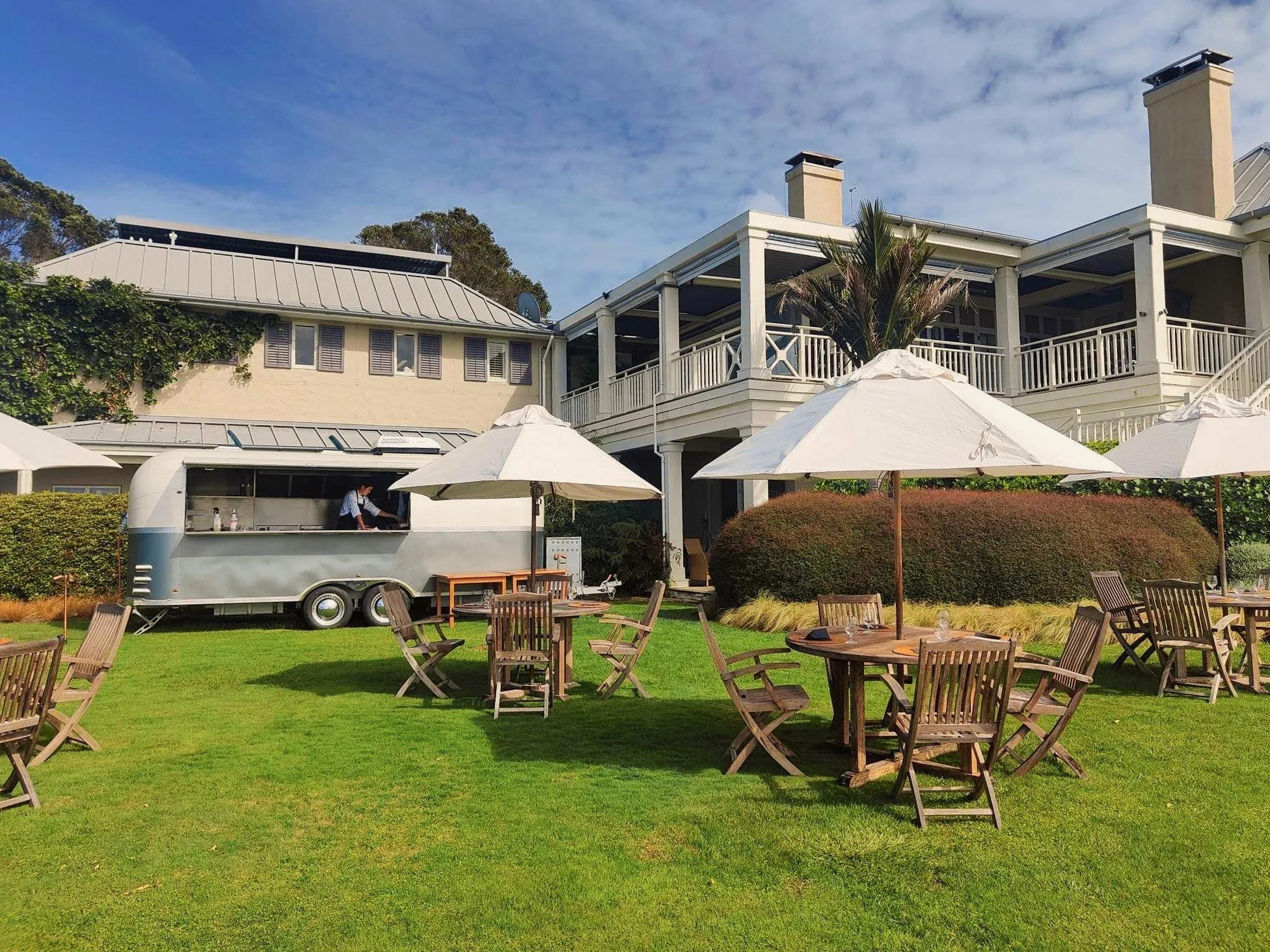 19th Tee food truck with outdoor tables with white umbrellas on the lawn in the foreground and the lodge in the background.