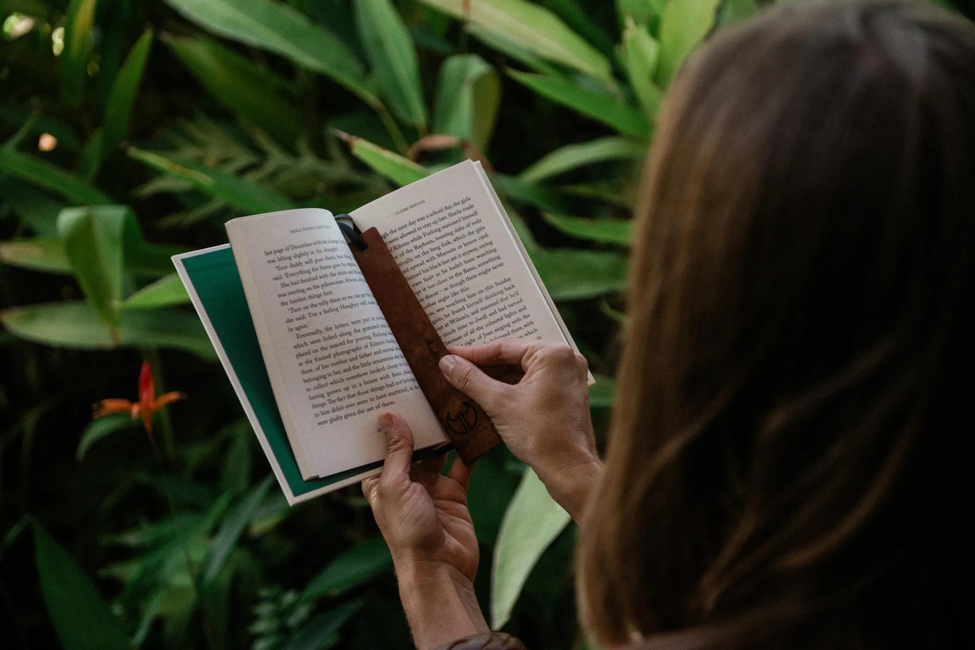 Person holding an open book with a wooden bookmark, surrounded by green foliage.