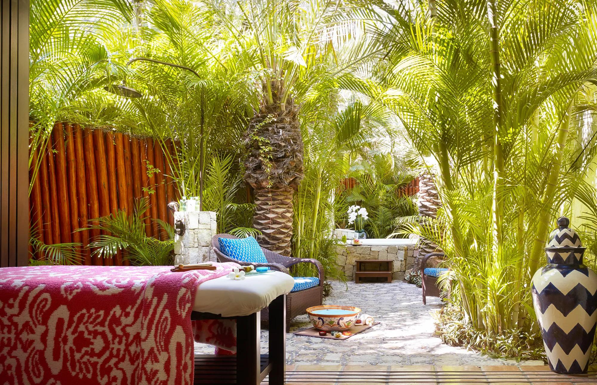 Outdoor spa area with a massage table, cushioned seating, tropical plants, and palm trees creating a serene and private atmosphere.