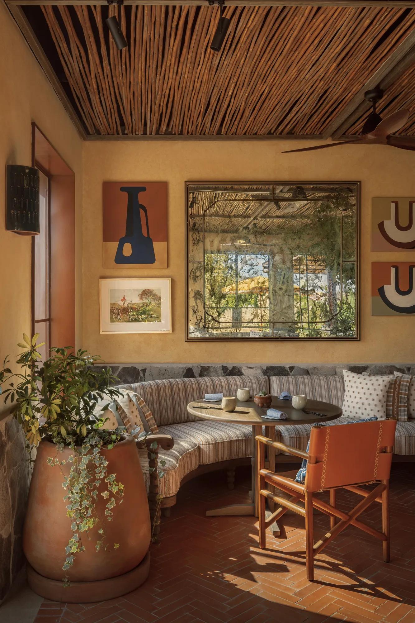 A round table set with napkins, cutlery, and glasses in a cozy corner. One chair has a woven backrest and an armchair is tucked against the wall adorned with framed paintings, an antique mirror, and a potted plant at the end of the armchair. 