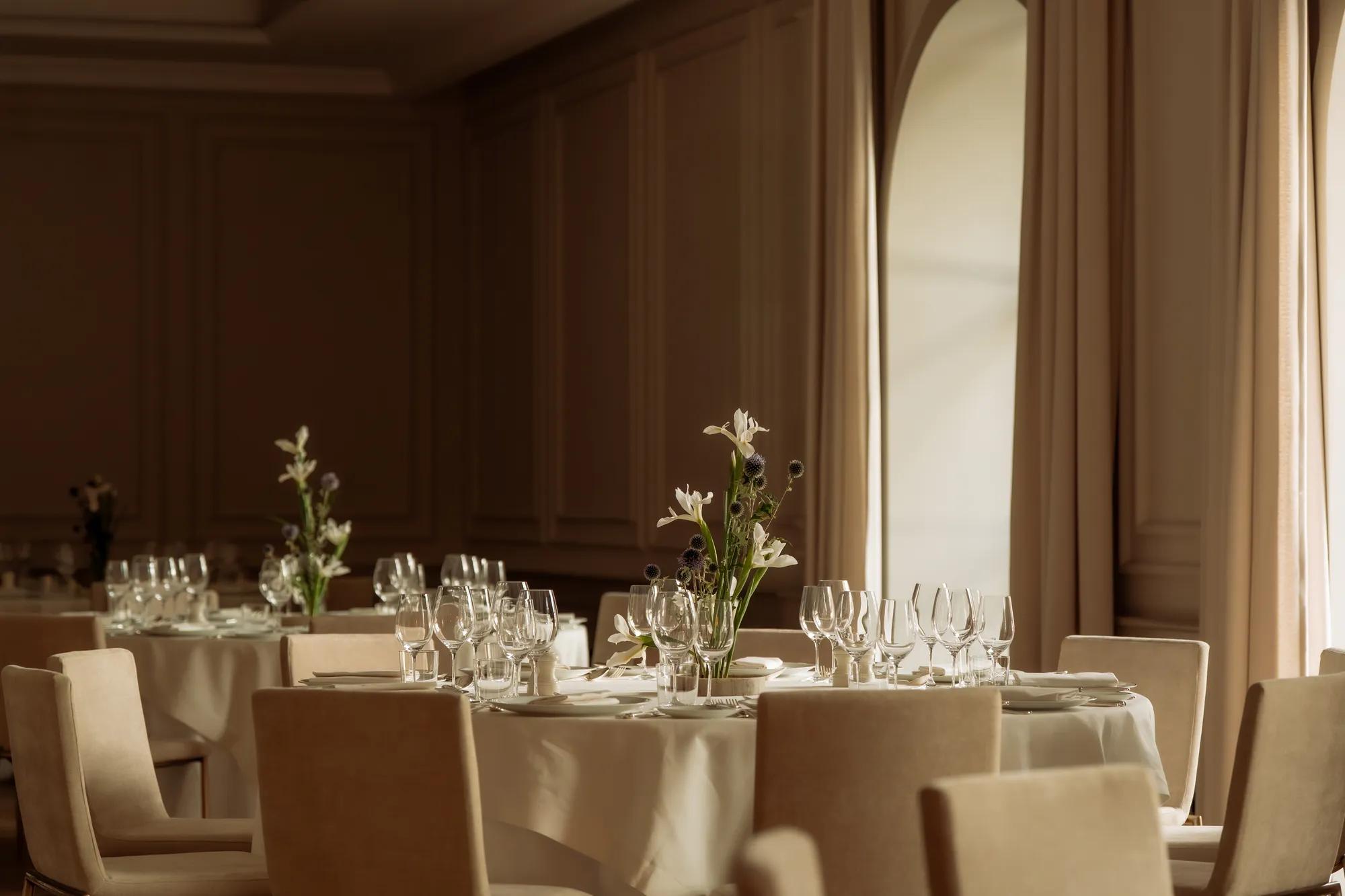 Wood panelled room with arched windows and round tables draped in white linens, decorated with glassware, china and floral arrangements. 
