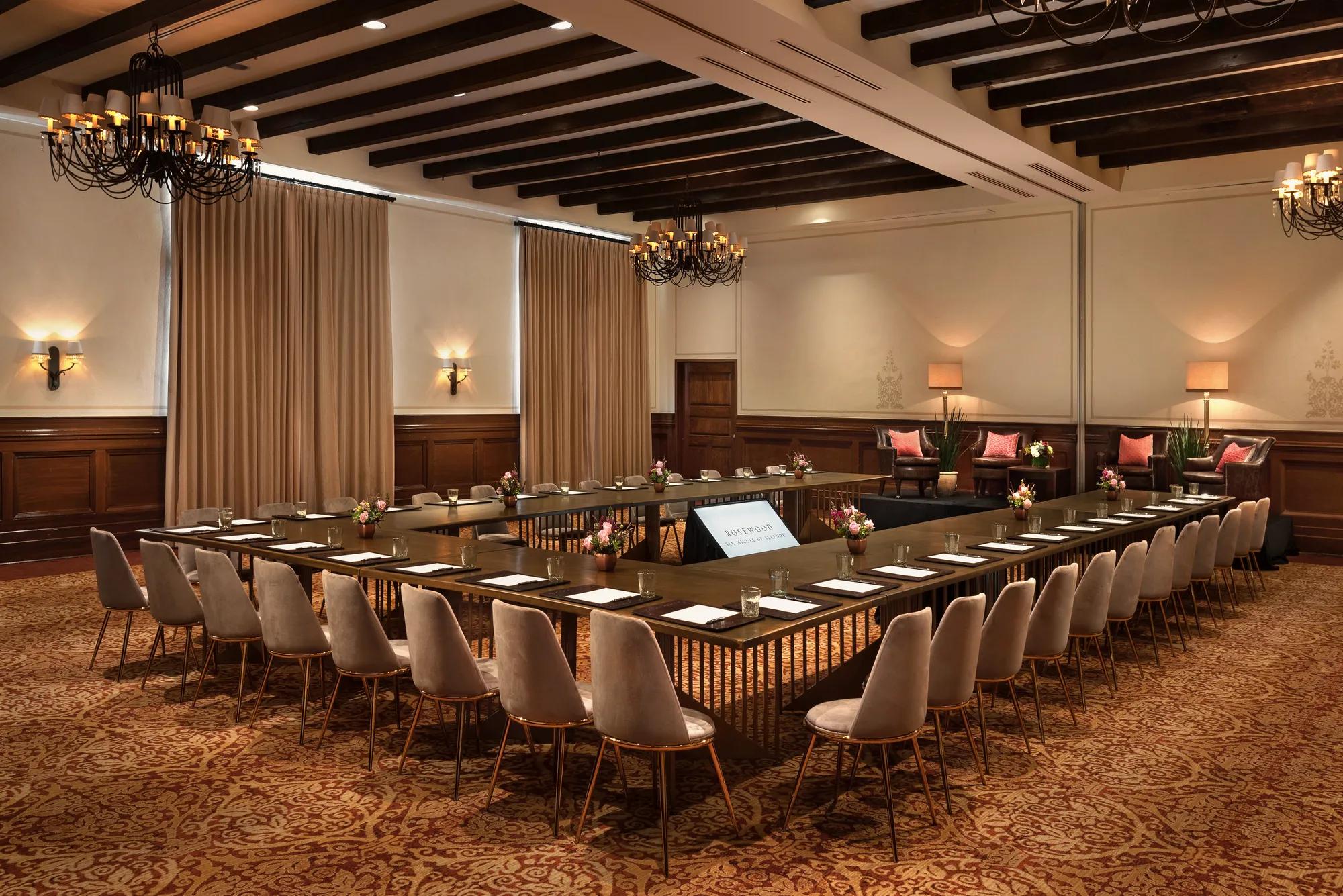 Ballroom set up in U-shape with carpeted floor, chairs and writing materials at each place, two large curtained windows on the left, and a speaker area with four armchairs at the front.
