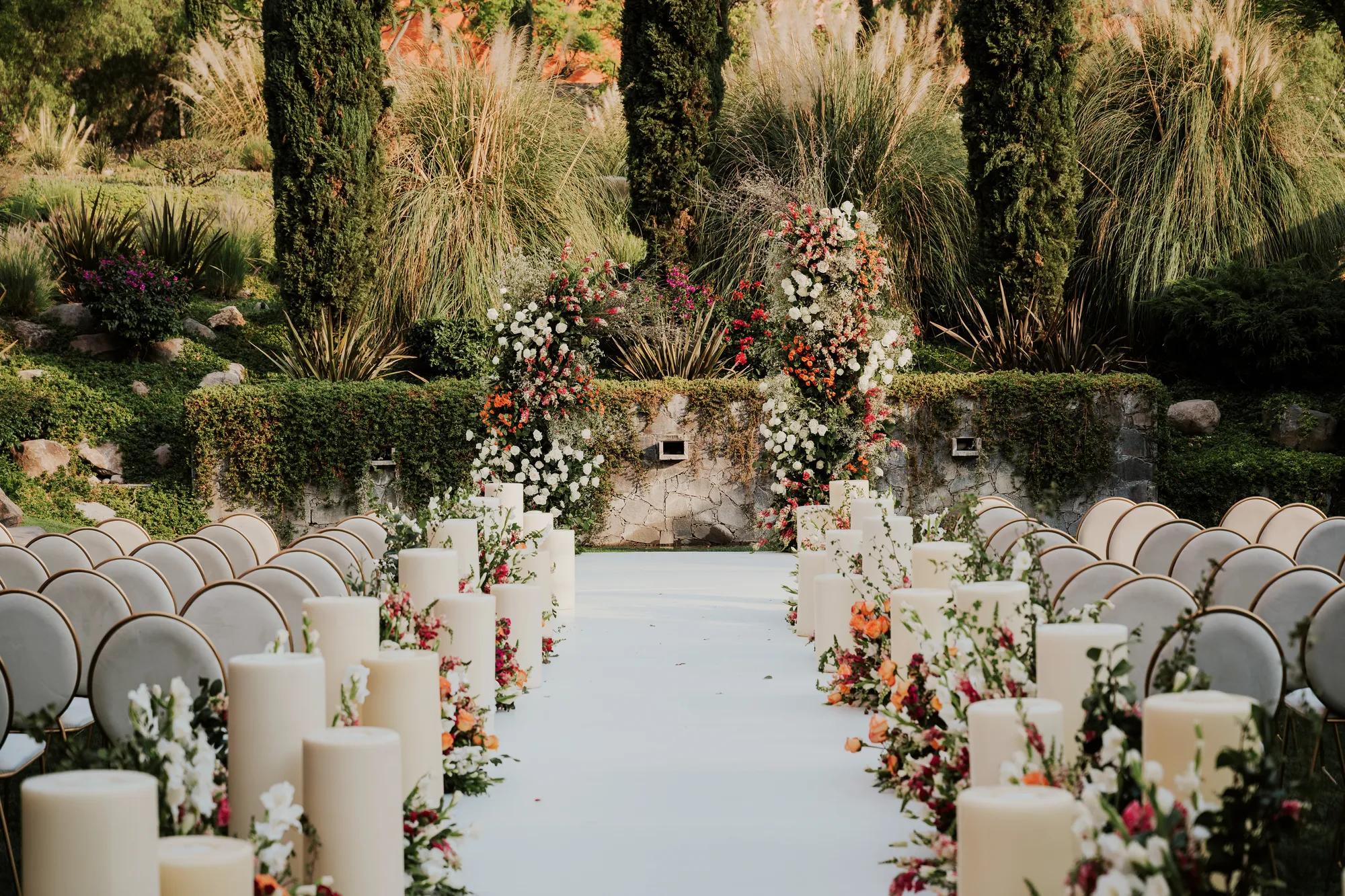 Wedding ceremony setup in a garden featuring a white aisle, rows of chairs on each side, and a floral arch at the end, surrounded by greenery.