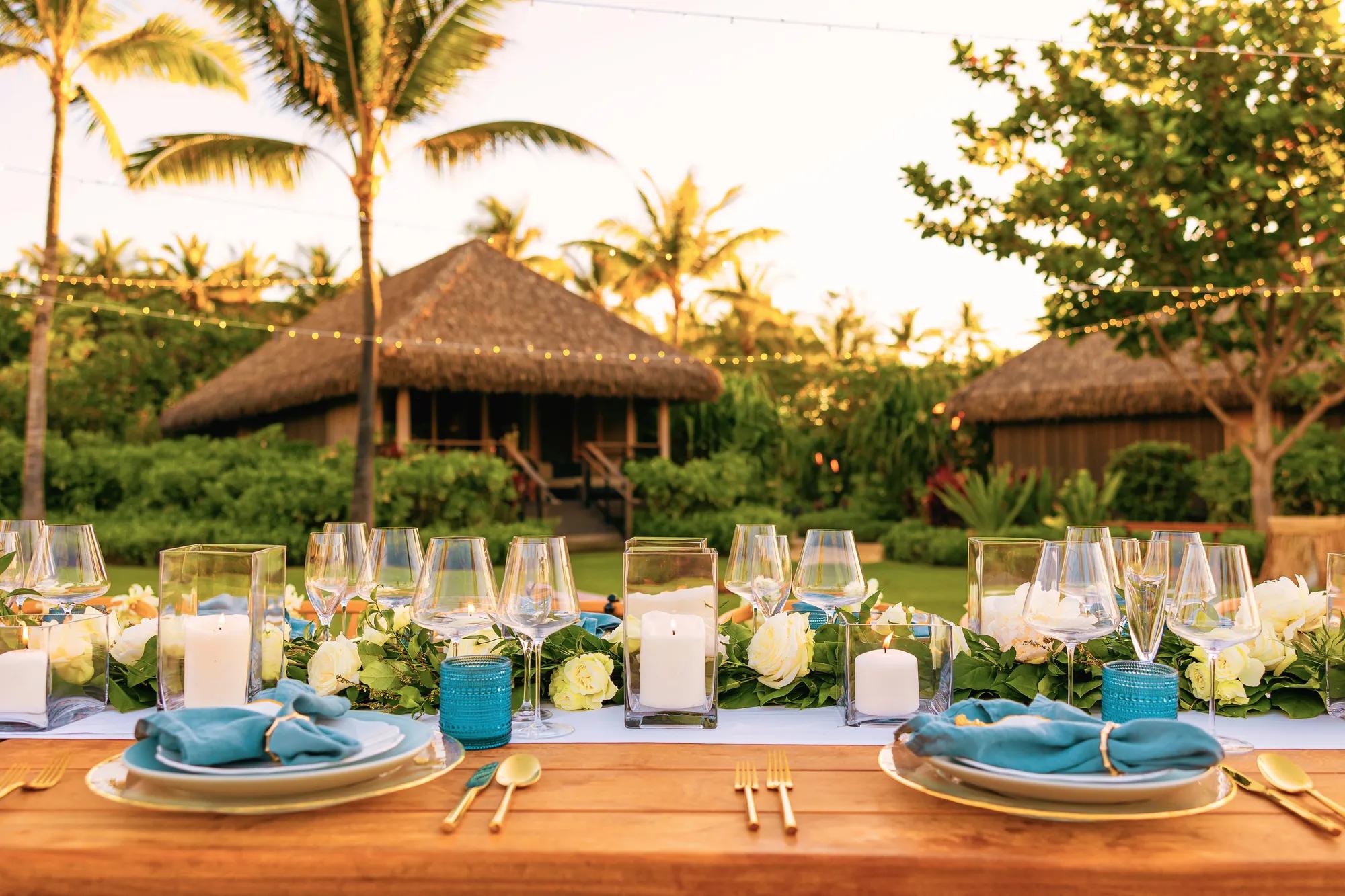 A horizontal tabletop set for an event on a lawn with thatched roof bungalows in the background