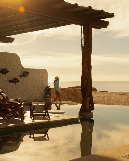 A luxury villa with an open-air patio and pool, featuring lounge chairs, a shaded daybed, and indoor living spaces visible through wide open doors, with bougainvillea adding color to the sunlit setting.