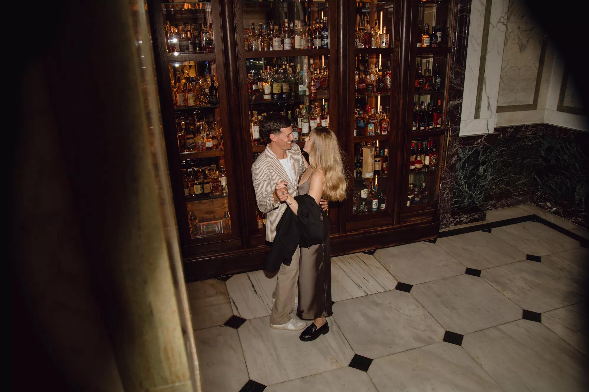 Couple dancing together in an intimate whisky room at Rosewood London, surrounded by marble and glass fronted spirits cabinets.