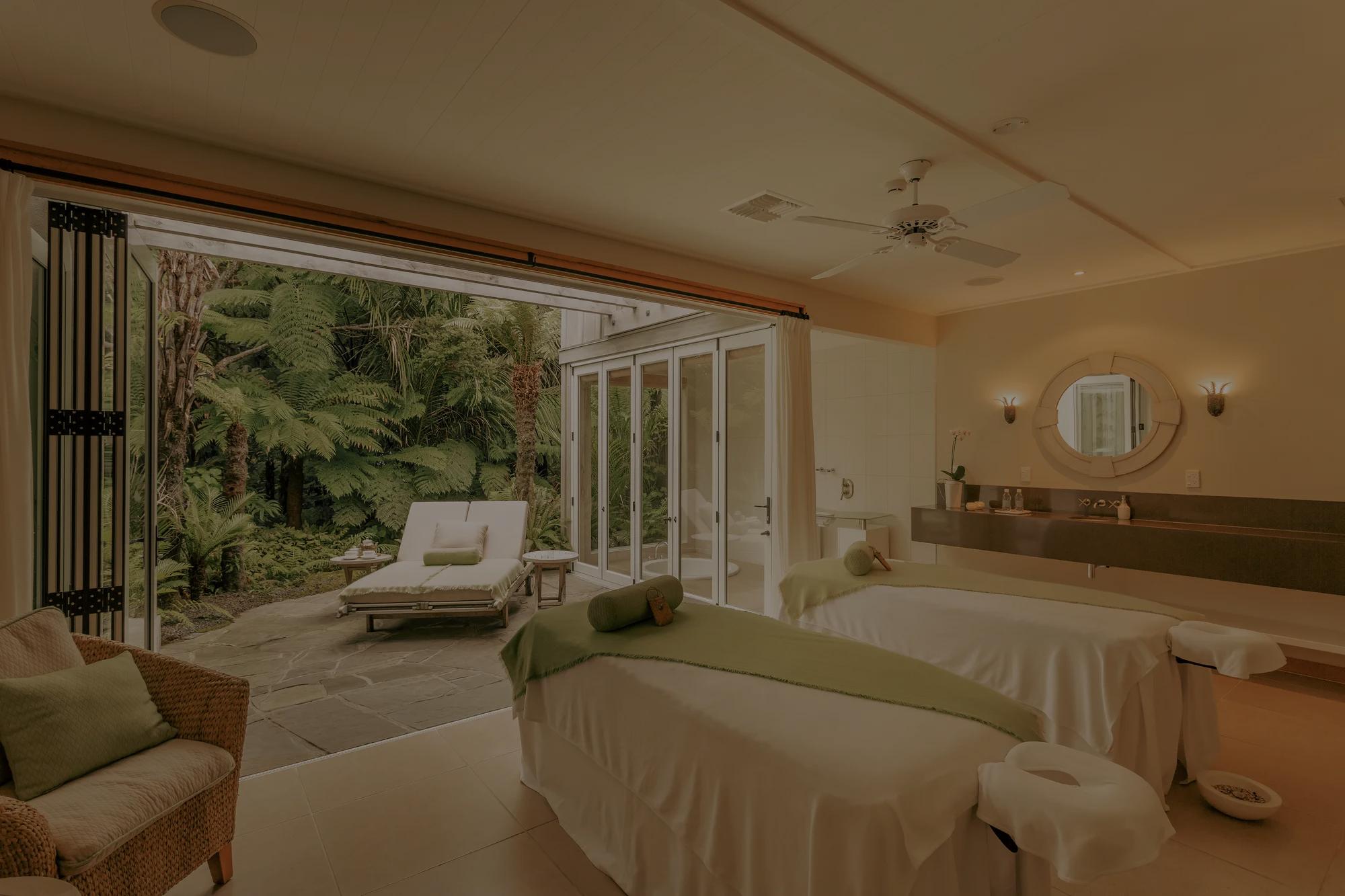 Couples spa room in a luxury hotel with two massage beds, an armchair and doors open to a stone terrace surrounded by native bush.