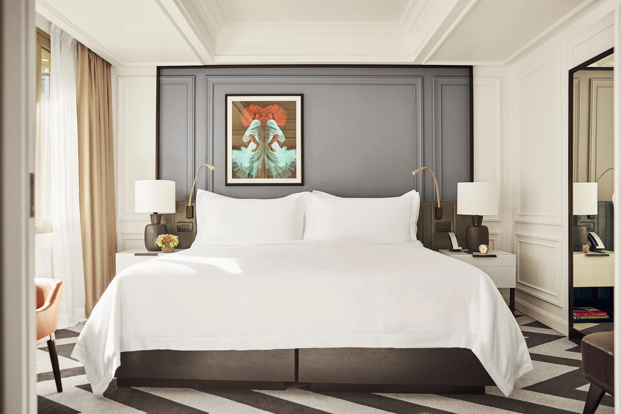 A hotel room with a large bed, lamps, a vertical mirror, and a window.