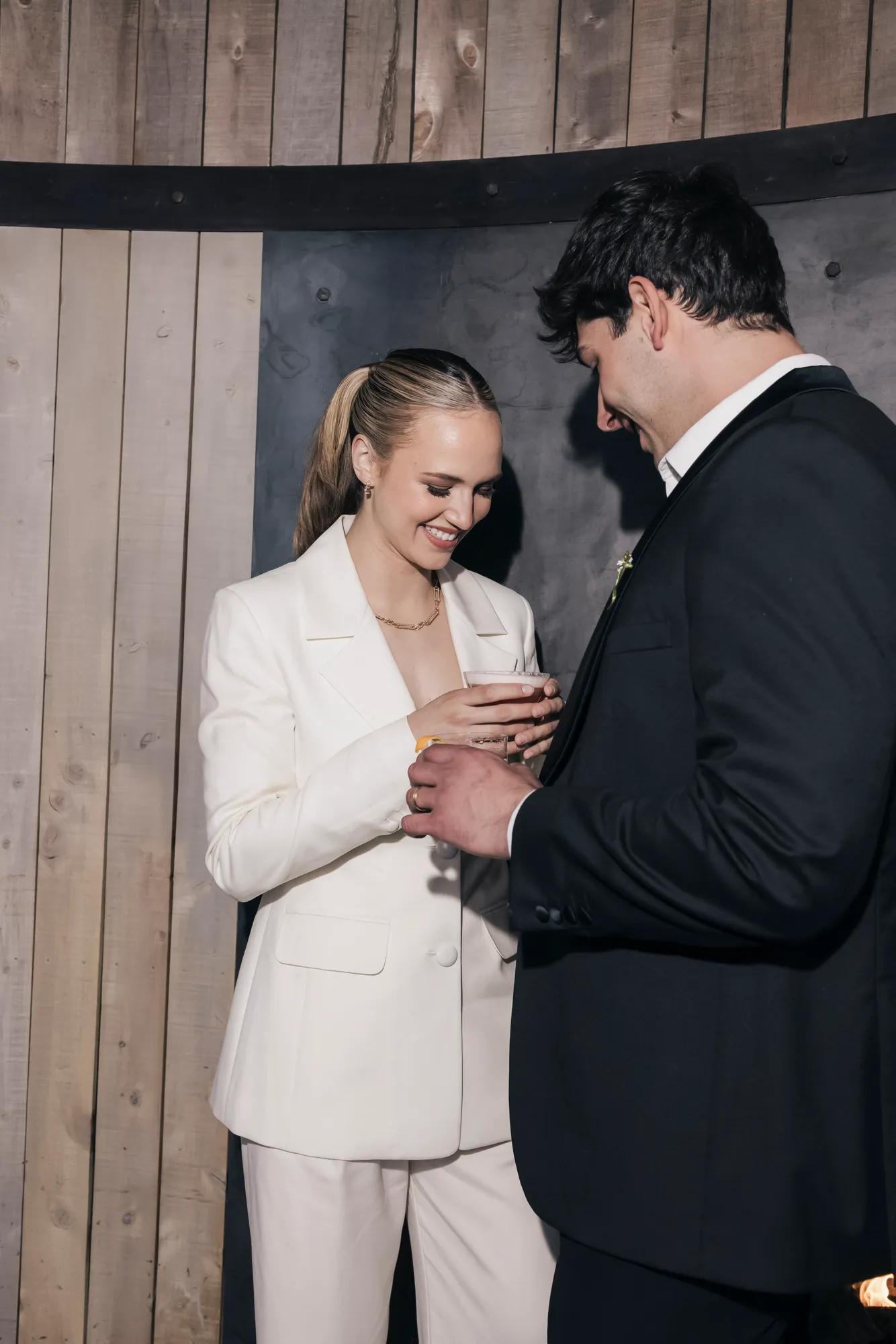 Bride in the white stylish tux holding a cocktail stands next to a groom in a cosy wood and steel clad room.