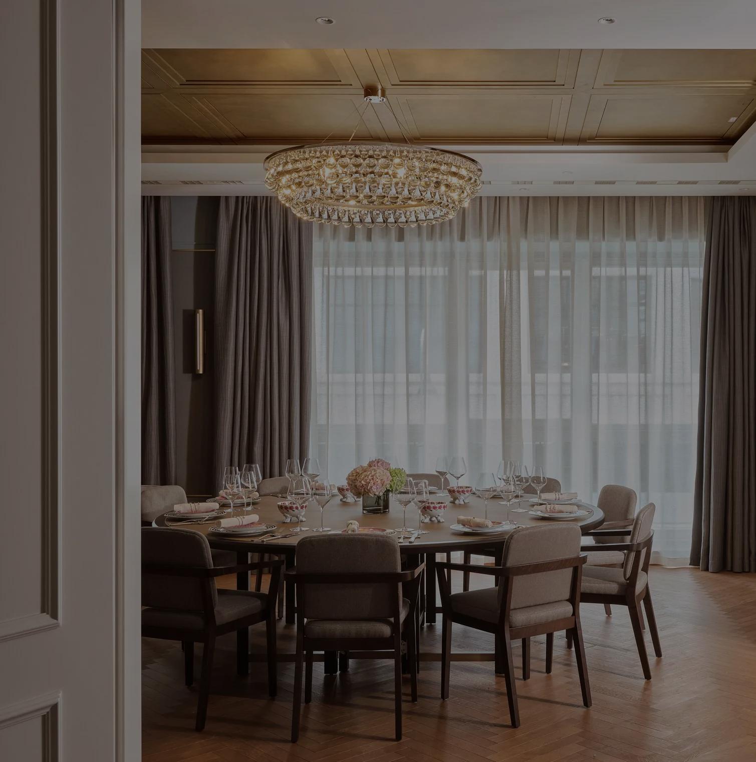 Elegant private event space at Rosewood Vienna featuring a round dining table set with glassware, fresh florals, grey upholstered chairs, floor-to-ceiling drapes, and a crystal chandelier.