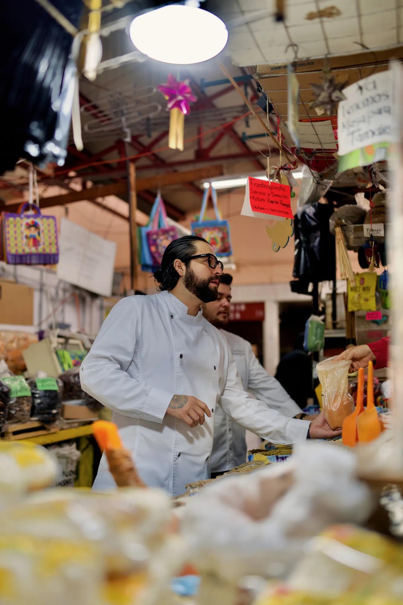Chef selecting fresh produce at a lively market, with vibrant bags hanging in the background.