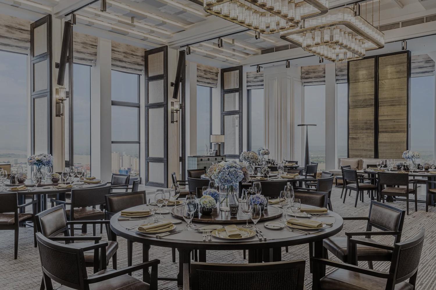 A luxurious dining room with large windows, round tables set with elegant tableware, and floral centerpieces, featuring modern chandeliers and a panoramic city view.