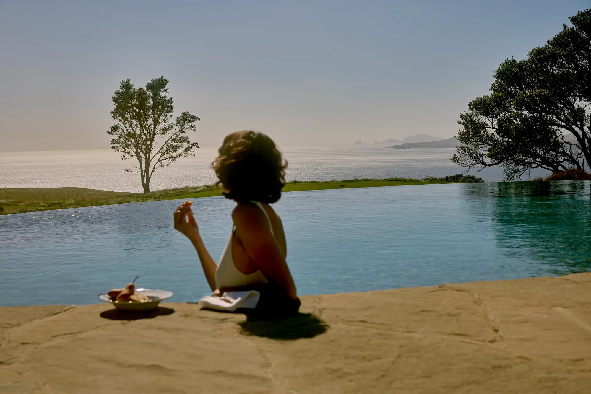 Woman leans on the edge of an infinity pool eating a slice of Pear as she looks out at views of the golf course and the Pacific Ocean.