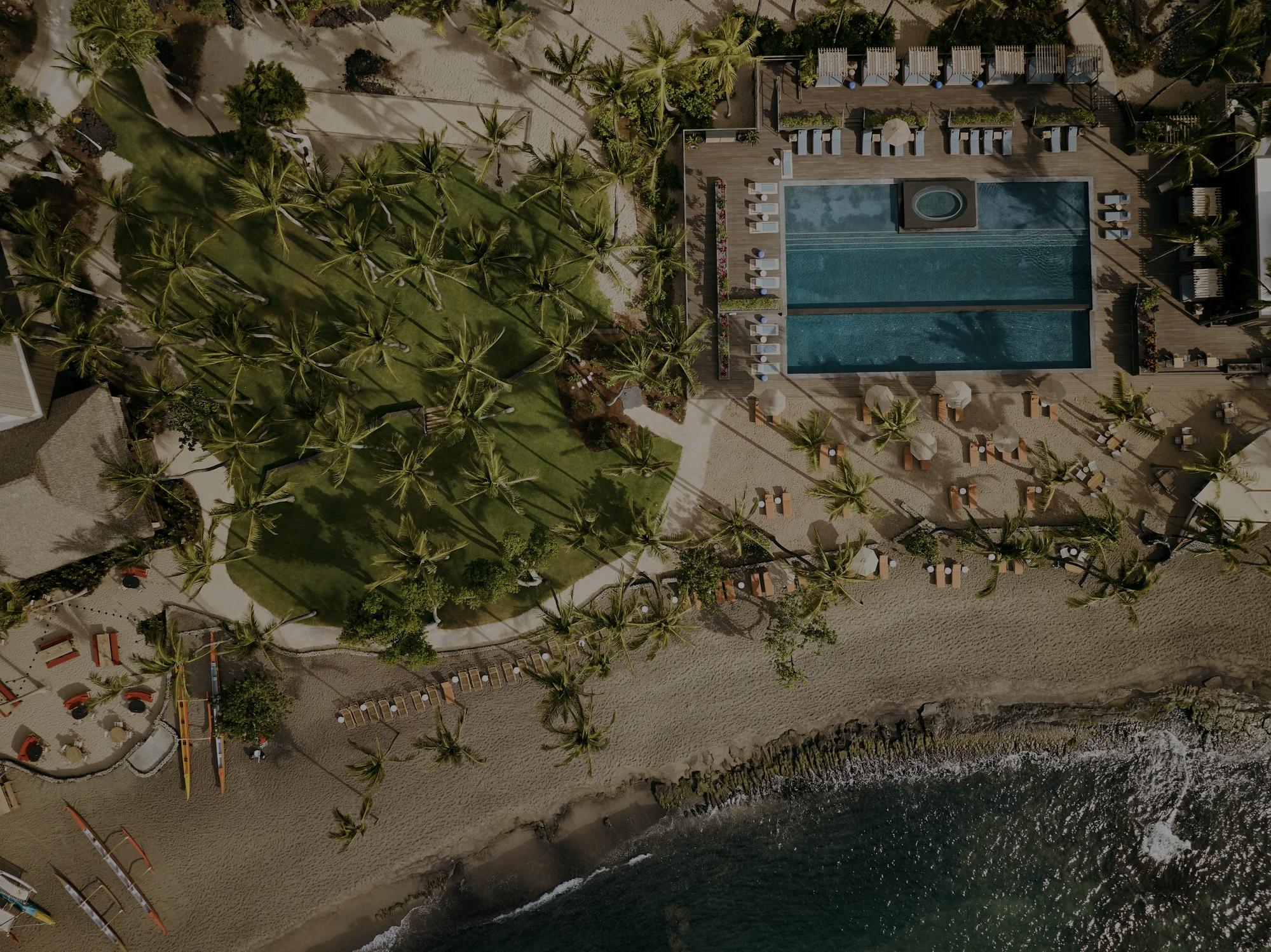 Aerial drone shot of vibrant green lawn next to sparkling blue pool and accross from the ocean with palms trees dotted accross the landscape in addition to beach loungers sprawled accross the beach and near the pool