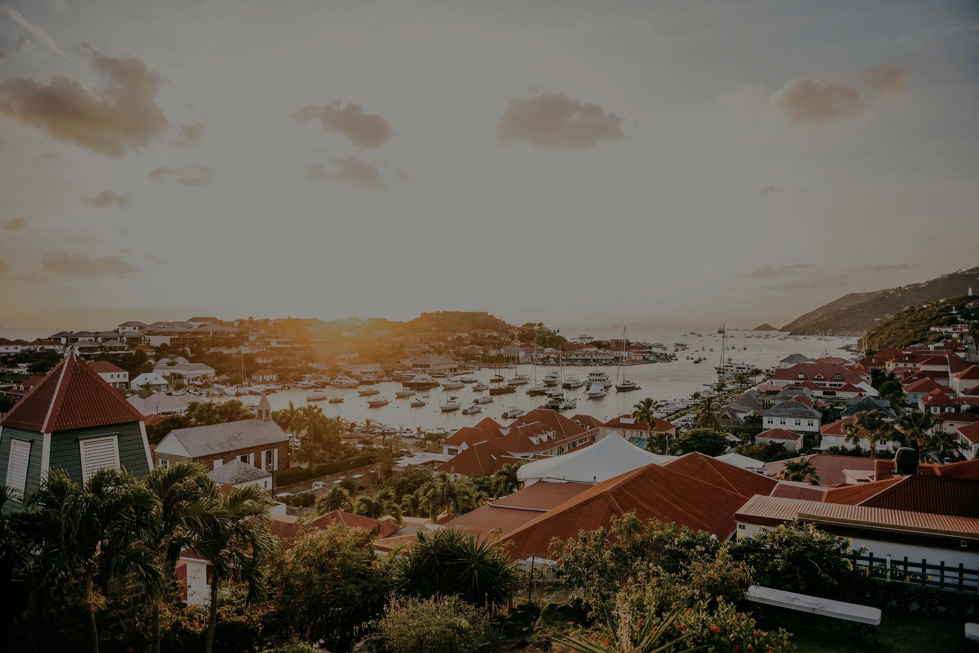 Evening view of St. Barth Beach with clear water, resort buildings, a mountain, and thatched umbrellas along the beach