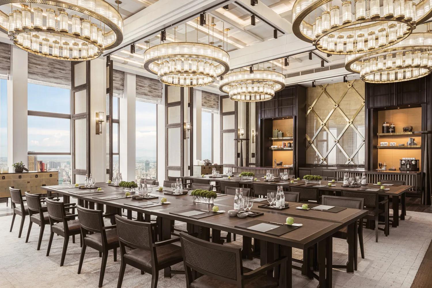 A large, elegant dining room with U-shaped tables set with placemats, glassware, and green plant centerpieces, surrounded by chairs and illuminated by ornate chandeliers, with city views through large windows.