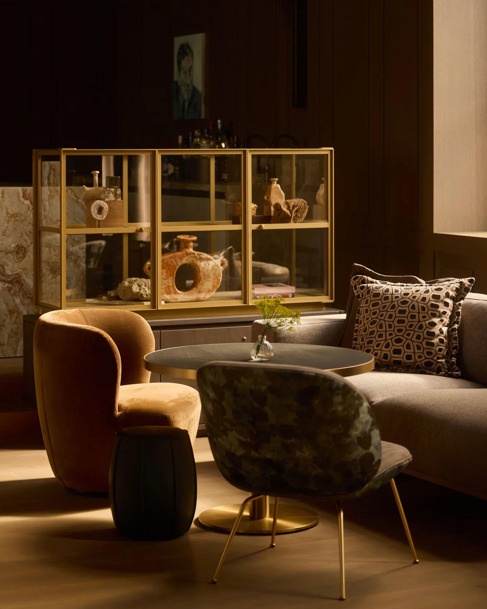 A room with jewel toned velvet sofas and armchairs next to a coffee table. Art and a vitrine cabinet of decorations is in the background. 
