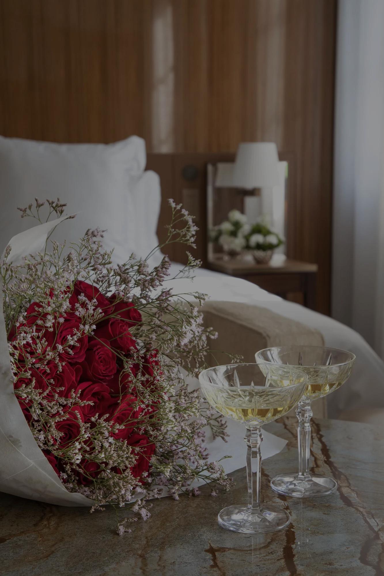 - Romantic setup at Rosewood São Paulo with a bouquet of red roses and two glasses of champagne on a marble table beside a bed with crisp white linens.